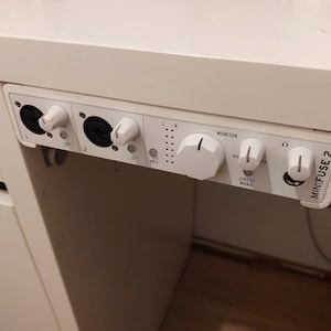 May include: A white Minifuse 2 audio interface mounted under a desk. The device features two XLR input ports with gain knobs, a central monitor volume dial, a direct mono switch, and a headphone jack with a dedicated volume control knob.