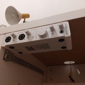 Könnte beinhalten: Ein weißes Minifuse 2 Audio-Interface, das unter einem Schreibtisch montiert ist. Das Gerät verfügt über zwei XLR/TRS-Kombi-Eingänge, mehrere Drehregler für Gain und Monitor-Pegel, einen Kopfhöreranschluss und Kontrollleuchten. Der Text Minifuse 2 steht an der Seite.