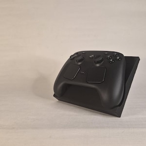 Pu&ograve; includere: Un controller per videogiochi nero opaco appoggiato su un supporto espositivo inclinato nero coordinato. Il controller presenta due levette analogiche, un pad direzionale, quattro pulsanti azione etichettati A, B, X e Y e due ampi touchpad rettangolari.