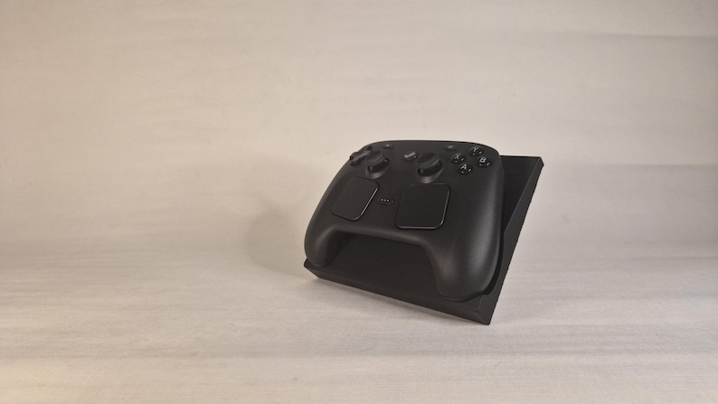 Pu&ograve; includere: Un controller per videogiochi nero opaco appoggiato su un supporto espositivo inclinato nero coordinato. Il controller presenta due levette analogiche, un pad direzionale, quattro pulsanti azione etichettati A, B, X e Y e due ampi touchpad rettangolari.