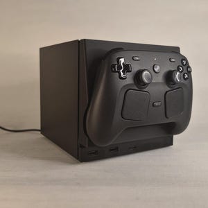 May include: A black gaming console dock featuring a matte finish with a black wireless controller mounted on the front. The console has a rectangular shape with visible ports at the base and a thin black power cable extending from the side of the unit.