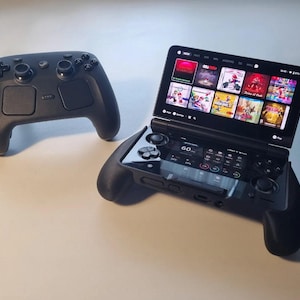 May include: A black handheld gaming console with a dual-screen display showing a game library and system settings, accompanied by a separate black wireless game controller featuring dual analogue sticks, a directional pad, and multiple action buttons.