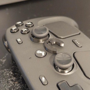 Könnte beinhalten: Ein mattschwarzer Gaming-Controller mit einem kreisförmigen Joystick-Zubehör, das neben dem linken Analogstick liegt. Der Controller verfügt über Standardtasten mit A-, B-, X-, Y-Beschriftung, eine Steam-Logo-Taste sowie Menü- und Navigationstasten.