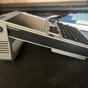 Könnte beinhalten: Ein tragbares Computergerät mit hellgrauem Gehäuse und schwarzer Tastatur, montiert auf einem passenden hellgrauen, verstellbaren Ständer. An der Seite befindet sich ein Anschluss mit der Aufschrift Expansion Port, der Bildschirm ist angewinkelt.