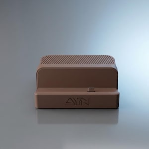 Op de afbeelding: Een bruin oplaadstation met een rechthoekige basis en de tekst AYN in reliëf op de voorzijde. Het apparaat bevat een centrale USB-C-connector en een bovenzijde met een reeks parallelle diagonale ventilatiesleuven voor warmteafvoer.