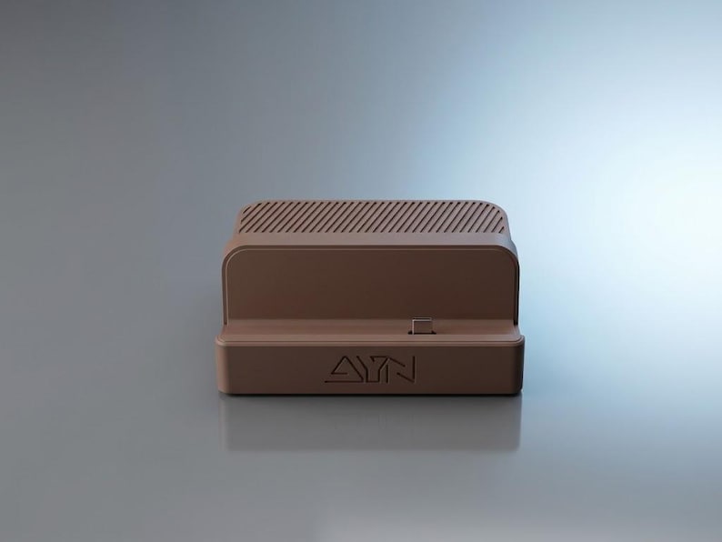 Op de afbeelding: Een bruin oplaadstation met een rechthoekige basis en de tekst AYN in reli&euml;f op de voorzijde. Het apparaat bevat een centrale USB-C-connector en een bovenzijde met een reeks parallelle diagonale ventilatiesleuven voor warmteafvoer.