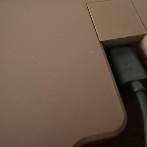 Op de afbeelding: Een matbruin oppervlak van een elektronisch apparaat met een verzonken poortgedeelte waar een zwarte oplaadkabel in is gestoken. De connector van de kabel toont het in reli&euml;f gemaakte logi-logo, wat duidt op een verbinding.