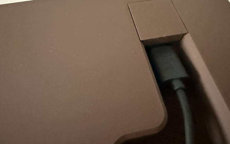 Op de afbeelding: Een matbruin oppervlak van een elektronisch apparaat met een verzonken poortgedeelte waar een zwarte oplaadkabel in is gestoken. De connector van de kabel toont het in reli&euml;f gemaakte logi-logo, wat duidt op een verbinding.