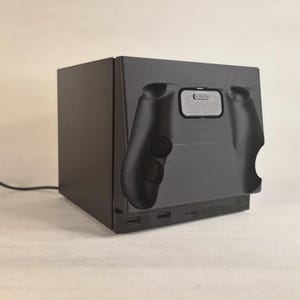 Pu&ograve; includere: Una base di ricarica elettronica nera di forma rettangolare con la sagoma stampata di un controller per videogiochi sul pannello frontale. Il dispositivo include due porte USB anteriori e un sottile cavo di alimentazione nero sul lato.
