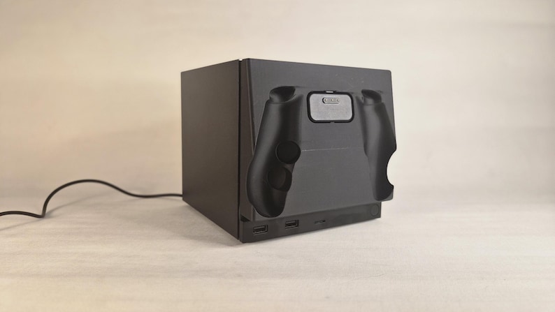 Pu&ograve; includere: Una base di ricarica elettronica nera di forma rettangolare con la sagoma stampata di un controller per videogiochi sul pannello frontale. Il dispositivo include due porte USB anteriori e un sottile cavo di alimentazione nero sul lato.