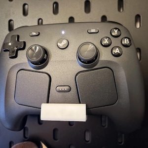 以下が含まれることがあります： 中央にSteamロゴボタン、2つのアナログスティック、十字キー、A・B・X・Yと刻印された4つのアクションボタン、2つの大きなタッチパッドを備えたマットブラックのビデオゲームコントローラー。底部には小さな白い長方形のスタンドが取り付けられています。