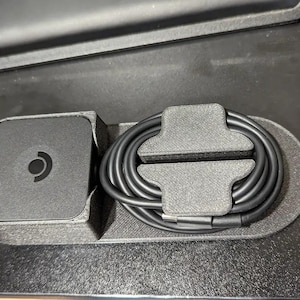 May include: A black power adapter featuring a circular logo with a central dot and a curved line, housed in a textured gray plastic organizer tray alongside a coiled black charging cable secured by a matching gray plastic cable management clip.
