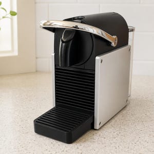 May include: A compact espresso machine featuring a matte black body with silver side panels, a chrome-finished lever, a black drip tray with horizontal slats, and a clear water reservoir attached to the back of the unit for brewing coffee.
