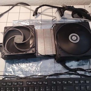May include: Two black computer fans with silver heat sinks are connected to a power supply and a keyboard. The fans are square with a central fan blade design. Wires and cables are connected to the fans.