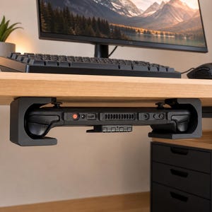 May include: A black under-desk mount securely holding a handheld gaming console. The mount is attached to the underside of a light wood desk, featuring a U-shaped design that cradles the device, keeping the console's top buttons and vents visible.
