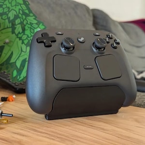 May include: A matte black wireless video game controller featuring a directional pad, two analog sticks, four action buttons, two large touchpads, and a central button with a Steam logo, resting on a black stand on a wooden surface.