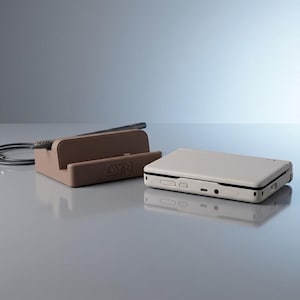 Op de afbeelding: Een beige handheld spelcomputer met een rechthoekige behuizing en een bruin oplaadstation met het AYN-logo. De console heeft zichtbare poorten en knoppen aan de zijkant, terwijl het station een zwarte oplaadkabel aan de achterzijde heeft.