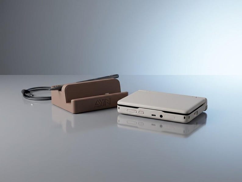Op de afbeelding: Een beige handheld spelcomputer met een rechthoekige behuizing en een bruin oplaadstation met het AYN-logo. De console heeft zichtbare poorten en knoppen aan de zijkant, terwijl het station een zwarte oplaadkabel aan de achterzijde heeft.
