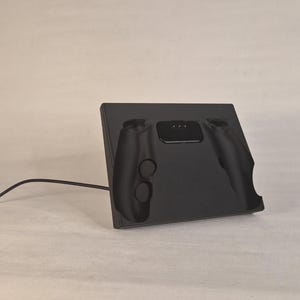 Pu&ograve; includere: Una base di ricarica nero opaco per controller di videogiochi. La base rettangolare presenta un incavo sagomato per alloggiare saldamente un controller, con una porta di connessione centrale e un cavo di alimentazione nero che si estende a sinistra.