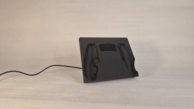 Pu&ograve; includere: Una base di ricarica nero opaco per controller di videogiochi. La base rettangolare presenta un incavo sagomato per alloggiare saldamente un controller, con una porta di connessione centrale e un cavo di alimentazione nero che si estende a sinistra.