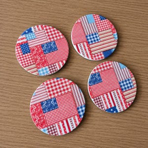 May include: Four round coasters with a patchwork design in red, white, and blue. The coasters feature various patterns, including gingham, stripes, and paisley, creating a patriotic theme. The coasters are arranged on a wooden surface.
