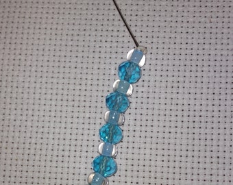 May include: A close-up of a needle threaded with blue and clear glass beads and a turquoise bead. The needle is resting on a white fabric background.