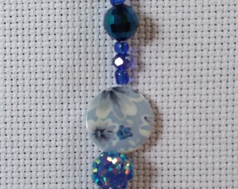 Könnte beinhalten: Ein Perlenanhänger mit einem blauen Metallring. Der Anhänger besteht aus einer Reihe blauer Perlen, einer Scheibe mit Blumenmuster, einem glitzernden blauen Ball und einer silbernen Filigrankugel. Die Perlen variieren in Größe und Farbton.
