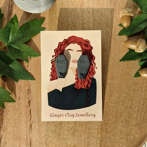 May include: A pair of gray clay earrings shaped like coffins with the word "RIP" on them. The earrings are shown on a model with red hair and a black shirt. The image also includes the text "Ginger Clay Jewellery".