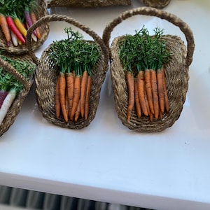 Könnte beinhalten: Kleine geflochtene Körbe gefüllt mit bunten Karotten und grünem Kraut. Die Karotten sind orange, gelb, rot und lila. Die Körbe haben einen Henkel und sind auf einer weißen Oberfläche angeordnet.