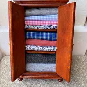 May include: A small, wooden wardrobe with open doors, displaying folded fabrics in various colours and patterns. The wardrobe is a rich brown colour and has small gold-coloured knobs. The fabrics include grey, pink, blue, and floral prints.