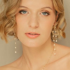 Puede incluir: Una mujer con cabello rubio y ojos azules que lleva un collar y aretes de oro con perlas. El collar tiene un colgante de perla única, y los aretes presentan múltiples perlas colgantes. La mujer está mirando a la cámara.