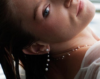 May include: A woman with wet hair and blue eyes wears a white sheer top with a delicate pearl necklace and pearl earrings.