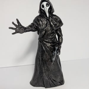 May include: A plague doctor figurine wearing a textured black hooded robe and a white bird-like mask. The figure has one arm extended forward with an open palm and holds a small silver-colored syringe in the other hand at its side.
