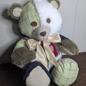 May include: A plush teddy bear constructed from patchwork fabric panels in shades of sage green plaid, white faux fur, and dark navy blue. The bear features a single brown button eye, a tan fabric nose, and a cream-colored satin ribbon tied around its neck.