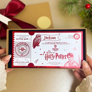 May include: A red and white Harry Potter-themed gift box with a Platform 9 3/4 ticket design. The box is held open, revealing the ticket design, with a red bow and gift box in the background. The text includes "Jackson" and "The Making of Harry Potter".