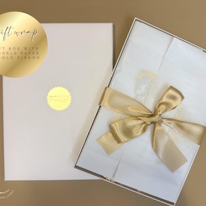 May include: A white gift box with a gold ribbon tied in a bow. The box is open, revealing white tissue paper. The lid of the box is closed. The text "Gift wrap" is on a gold circle.