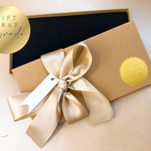 May include: A gift box with a black interior, wrapped with a cream-colored satin ribbon and a blank tag. The box is brown with a gold foil sticker. The words "GIFT WRAP upgrade" are in a gold circle.