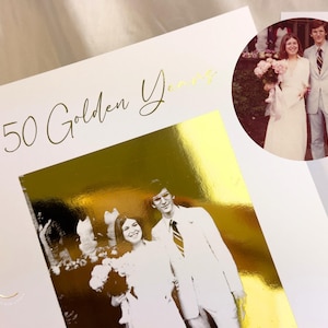May include: A white card with gold foil accents and the text "50 Golden Years" in elegant script. The card features a black and white photo of a couple, with a circular photo of the couple in the upper right corner. The card is a wedding anniversary theme.