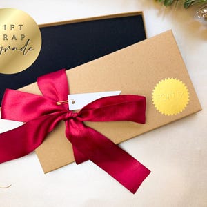 May include: A gift box with a black interior and a kraft paper exterior. The box is tied with a deep red satin ribbon and a small white tag. A gold foil sticker and a gold circle with the words "GIFT WRAP upgrade" are also visible.