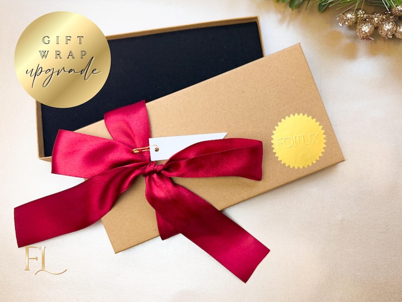 May include: A gift box with a black interior and a kraft paper exterior. The box is tied with a deep red satin ribbon and a small white tag. A gold foil sticker and a gold circle with the words "GIFT WRAP upgrade" are also visible.