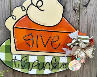 May include: A wooden door hanger shaped like a slice of pumpkin pie with a white whipped cream topping and cinnamon sticks on top. The pie crust is green and white checkered. The words "give thanks" are written in black on the pie. A small tag with the words "Our Tree Swing Studio 2023" is attached to the bottom right corner.