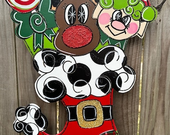 May include: A red, white, and black Santa boot door hanger with a green and red plaid pattern on the bottom. A reindeer with a red nose and a green elf with a red hat are peeking out from the top of the boot.