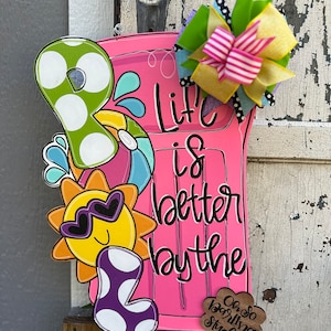 May include: Pink wooden door hanger with the text "Life is better by the" and a sun with sunglasses, a beach ball, and a purple polka dot pool float. The door hanger also has a small wooden tag with the text "Oh so darling studios".