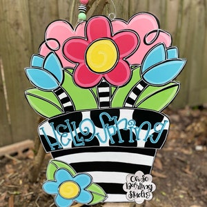 May include: A wooden door hanger with a black and white striped pot with the words "Hello Spring" in blue. The pot has pink, blue, and yellow flowers with green leaves. The bottom of the hanger has a white circle with the text "Oh So Darling Studio".