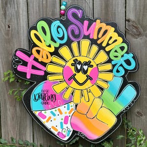 May include: A colorful wooden sign with the words "Hello Summer" in various bright colors. The sign features a smiling sun, a peace sign, and a section with sprinkles. The sign is designed to hang and has a wire and beaded accent.