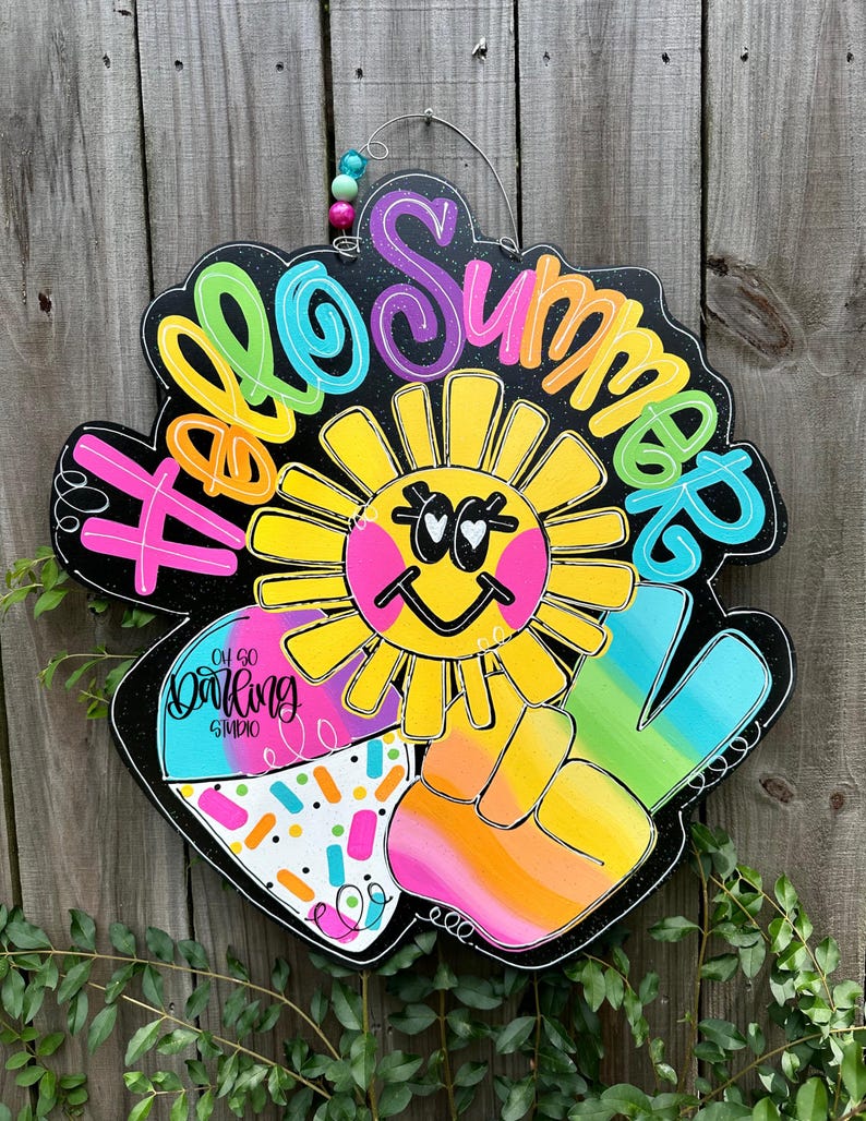 May include: A wooden sign with the words "Hello Summer" in colorful lettering. The sign features a smiling sun, a peace sign, and a section with sprinkles. The sign is black with a white border and a wire hanger.