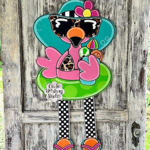 May include: A pink flamingo wearing sunglasses and a turquoise hat with a pink flower. The flamingo is holding a green and pink drink with an orange slice. The flamingo is sitting on a green inflatable ring. The flamingo has black and white polka dot legs and turquoise and orange flip flops. The text "Oh So Darling Studio" is on a white cloud.
