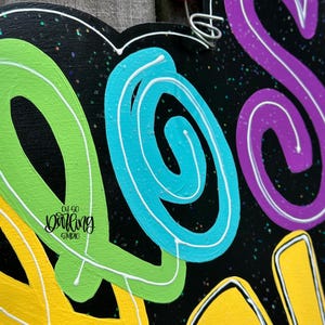May include: A black wooden sign with the word "SO" in bright, swirling colors: yellow, green, blue, and purple. The letters are outlined in white. The sign has a pink bead and wire for hanging. The words "Oh So Darling Studio" are also on the sign.