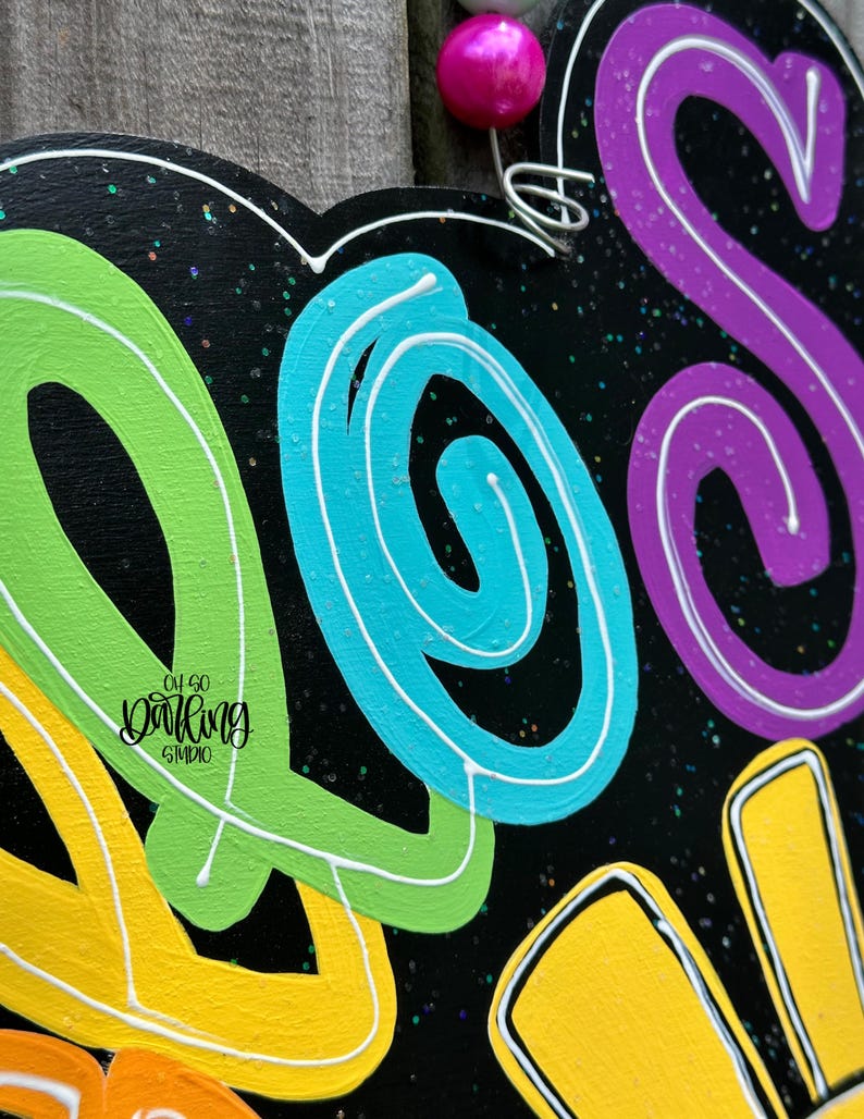 May include: A black wooden sign with the word "SO" in bright, swirling colors: yellow, green, blue, and purple. The letters are outlined in white. The sign has a pink bead and wire for hanging. The words "Oh So Darling Studio" are also on the sign.