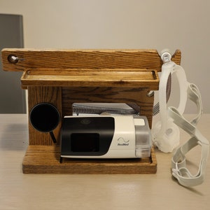 May include: A wooden stand with two shelves for a CPAP machine, mask, and other accessories. The stand is made of dark wood with a natural finish. The CPAP machine is white with a black display and the text "ResMed" on the front. The mask is white and has a clear tube.
